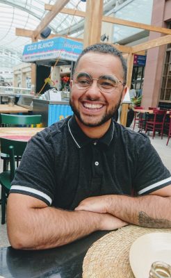 A smiling POC man sits at a table in a public space.
