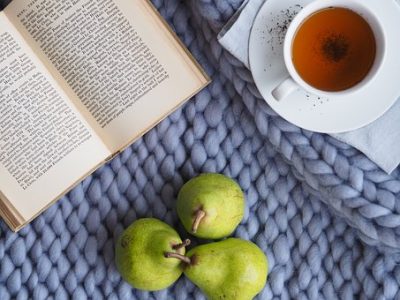 bird's eye view of an open book, a tea in a cup on a saucer, three pears, all placed on a blue-grey wool blanket