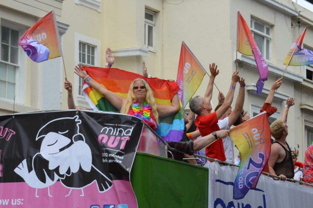 sue waving a rainbow flag on the top of the mindout bus at pride. other people are waving flags behind her and the mindout logo is displayed on the side of the bus.