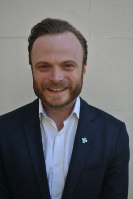 white man with light brown hair and a short beard smiling at the camera, wearing a white shirt and a blue blazer.
