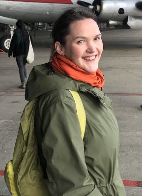 white woman with green jacket and backpack, orange scarf, smiling turning back at the camera. looks as though she is about to board an aeroplane.