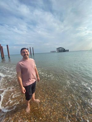A person standing in the shallow ocean, wearinga pink shirt and grey shorts, smiling into the camera