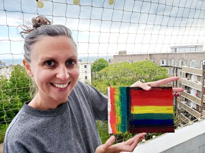 white woman, wearing a grey top, holding a pride flag she created.