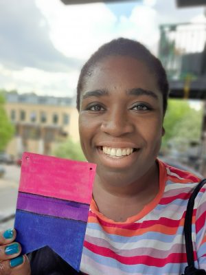 Black woman holding a bisexual flag which she created herself, she's smiling into the camera