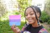 Brown woman holding a watercoloured bisexual flag, she's smiling into the camera