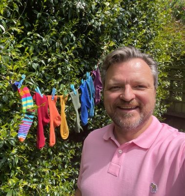 white man standing in front of colourful socks that resemble pride colours, he's wearing a pink shirt and has silver hair
