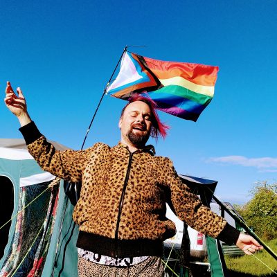 White person wearing a cheetah jacket, with the progressive Pride flag behind them