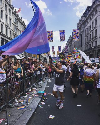 white man holding the trans flag, they're wearing a black top and white shorts