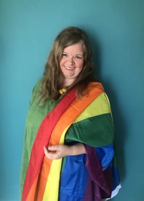 White woman with long brown hair, wearing the pride flag on her shoulder