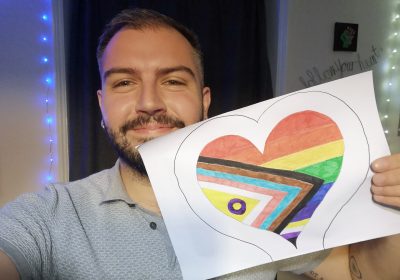 Fair skinned person holding the Intersex Inclusive flag that they drew themsleves, they are smiling into the camera