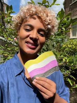 Curly haired person holding the non-binary flag that has been drawn, and they are smiling into the camera