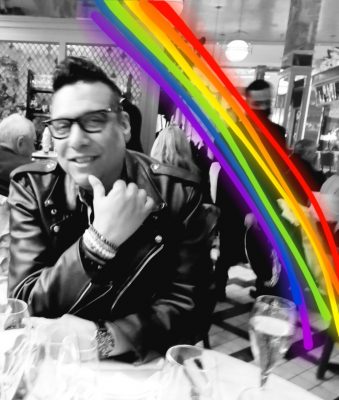 person sitting at a table, looking into the camera, wearing a black leather jacket. A rainbow has been drawn on the picture on the right hand corner