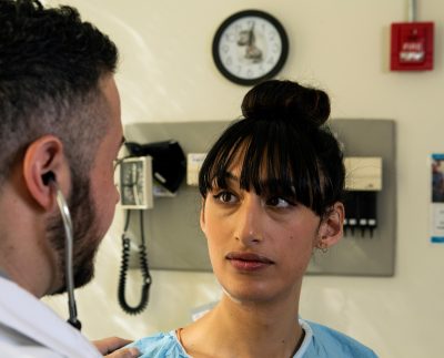 A transgender woman in a hospital gown having a conversation with a doctor, a transgender man.