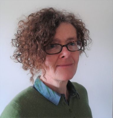 Helen wearing a green jumper with a blue colour, and glasses, turned to the side and looking at the camera