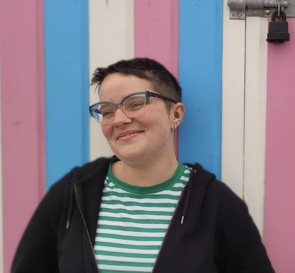 White appearing person smiling away from the camera, they are wearing a black hoodie and a green and white top and they are standing in front of a beach hut which has blue, pink and white stripes