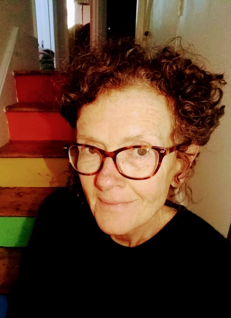White appearing women smiling into the camera, she has curly short hair and is sitting on rainbow steps