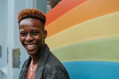 QTIPOC appearing person smiling into the camera, standing in front of painted wall of the pride colours
