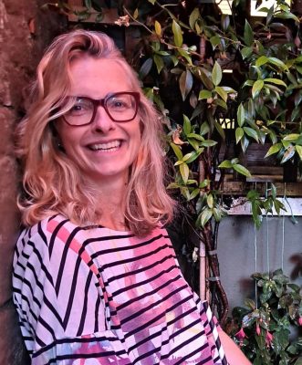 woman with long blonde hair wearing glasses smiling at the camera. sitting in front of some green foliage and the lighting of the photo is warm. she is wearing a stripey black and white shirt, and sitting adjacent to the cameraperson, so has her head turned to look at the camera.