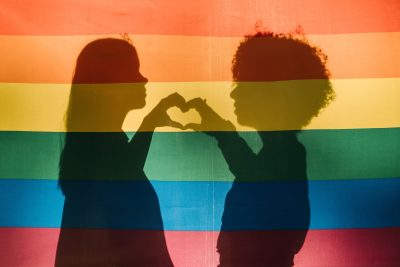 Two people standing behind the flag pride colours making a heart shape using thier hands