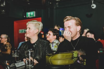 Two white appearing people watching the MindOut for the Laughs show, one person has their hand over their mouth and is squinting their eyes with laughter