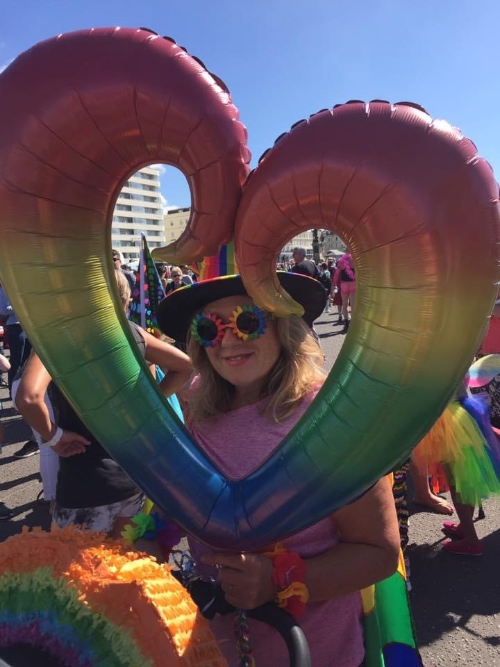 White appearing femme looking through a rainbow coloured heart balloon