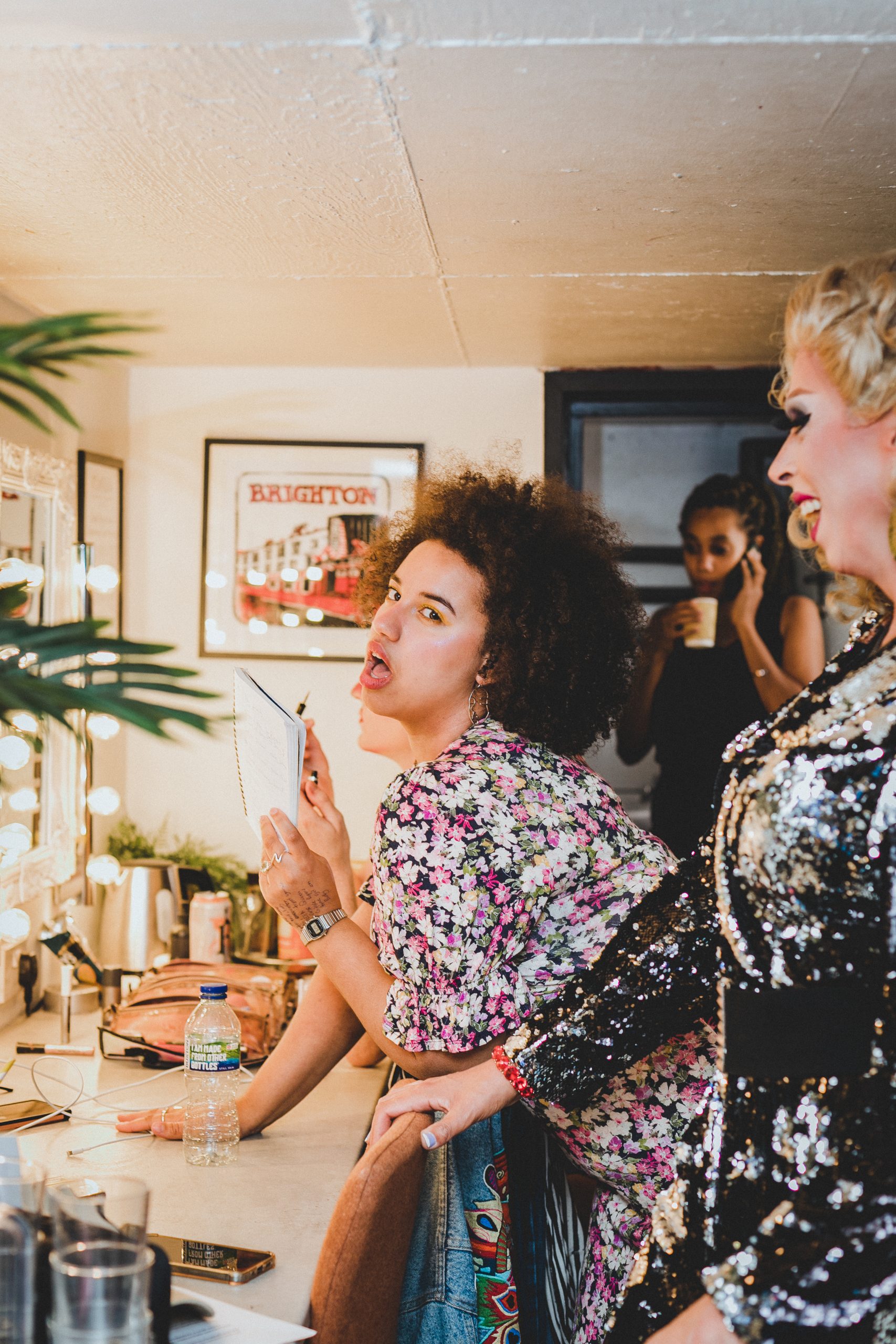 Emily Bampton, back stage posing at the camera, she has curly afro hair and wears a floral maxi dress. Ophelia Payne is stands next to Emily, smiling towards her. Kate Cheka, is stood in the background holding a cup and talking on the phone
