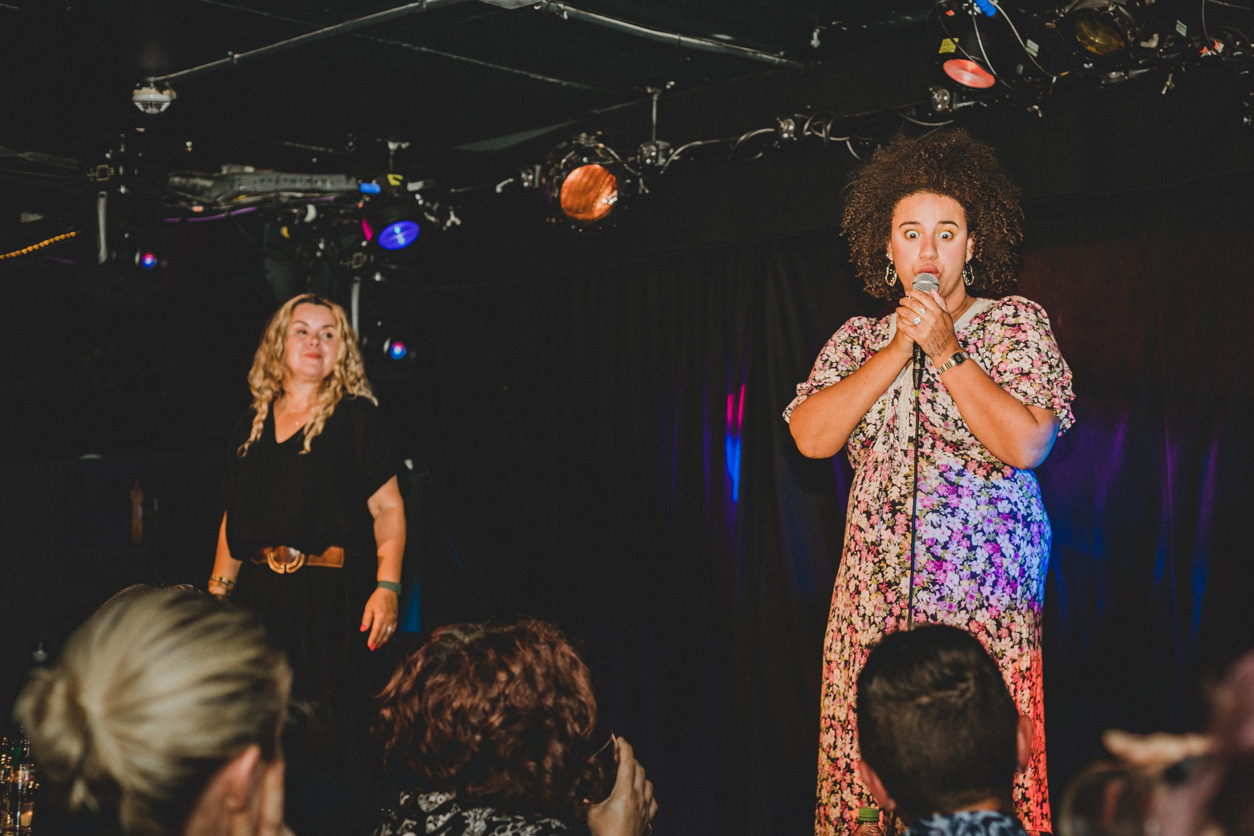 Emily Bampton, stand up comedian is on stage, wearing a floral maxi dress, she has curly afro hair and is holding the mic to her mouth. Standing to the right is Paula (BSL signer) who is wearing a black jumpsuit and has blonde long wavy hair