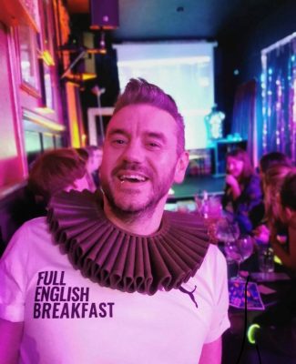 white appearing man sat in a bar, smiling into the camera, he has short brown hair and is wearing a long sleeve top that says 'full English breakfast'