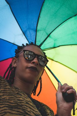 POC person wearing sun glasses and holding a rainbow umbrella looking down into the camera