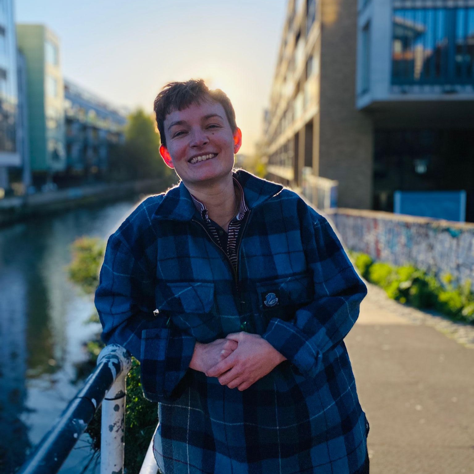 Tommy is leaning on a rail and smiling at the camera. They are standing on the bank of a canal and there is low sunlight shining behind them. They are wearing a blue flannel shirt.