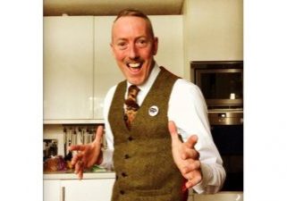 A smiling white man in a waistcoat and tie standing in a kitchen.