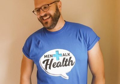 A white man with a short beard and glasses wearing a blue t-shirt that says 'Men Talk Health'. He is smiling and looking to the left of the frame.