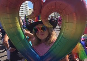 White appearing femme looking through a rainbow coloured heart balloon