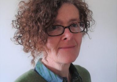 Helen wearing a green jumper with a blue colour, and glasses, turned to the side and looking at the camera