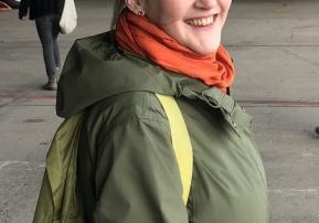 white woman with green jacket and backpack, orange scarf, smiling turning back at the camera. looks as though she is about to board an aeroplane.