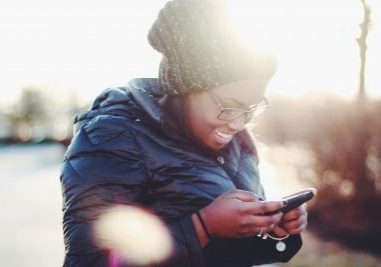 Black person with long hair and glasses smiling and looking down at their phone. they are outside and wearing a warm jacket and wooly hat.